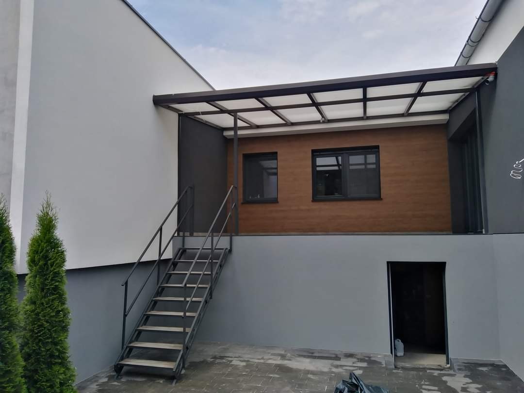 Nowoczesny dom z elewacją w odcieniach szarości i brązu, z metalowymi schodami prowadzącymi na zadaszony taras, widoczne dwa małe drzewka iglaste.