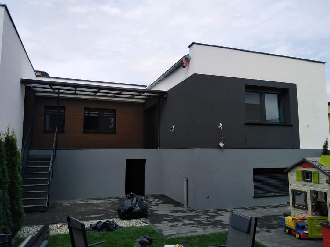 Nowoczesny dom z elewacją w odcieniach szarości i bieli, z drewnianym wykończeniem pod zadaszeniem tarasu i metalowymi schodami zewnętrznymi, widok z ogrodu.