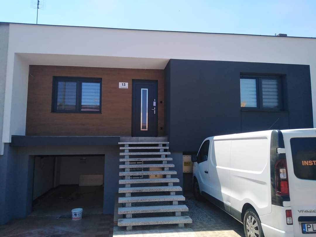 Nowoczesny dom z elewacją w odcieniach brązu i szarości, widoczne schody wejściowe, brama garażowa i zaparkowany biały samochód dostawczy.