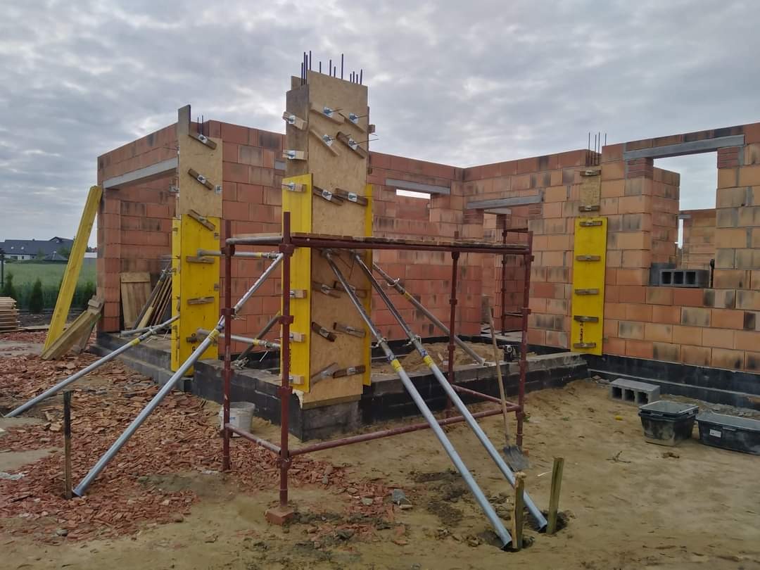 Wznoszenie ścian z czerwonej cegły domu jednorodzinnego, widoczne szalunki i rusztowania, teren budowy w trakcie prac.