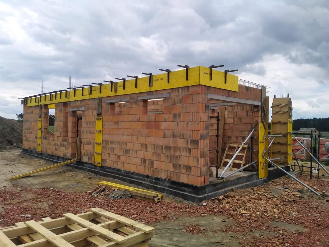 Wznoszenie ścian z czerwonej cegły na fundamencie, z użyciem żółtych szalunków i belek stropowych, widok na plac budowy w pochmurny dzień.