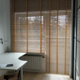 Jasne drewniane żaluzje poziome z szerokimi lamelami w pokoju z biurkiem, widoczne sznurki do regulacji, grzejnik pod oknem.