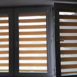 Okno z trzema skrzydłami, wyposażone w beżowo-białe rolety typu dzień i noc, jedno skrzydło otwarte.