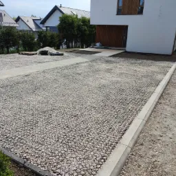 Utwardzony żwirem podjazd do nowoczesnego domu z betonową obwódką i ścieżką z kostki, widoczne przygotowane podłoże i paleta z materiałami budowlanymi.
