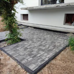 Ułożona szara kostka brukowa na posesji, z czarnym obrzeżem, rabata na drzewo wkomponowana w nawierzchnię, widoczny fragment elewacji budynku z oknami piwnicznymi.