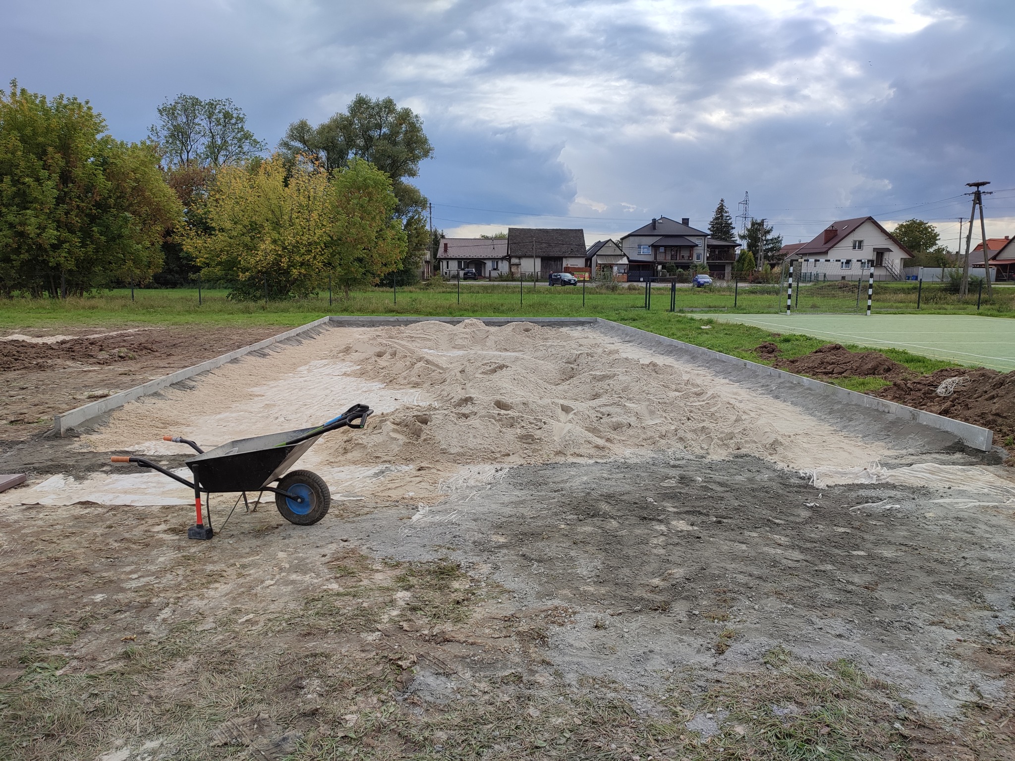 Przygotowany teren pod budowę z widoczną warstwą piasku otoczoną betonową obwódką, taczka z niebieskim kołem stoi obok, w tle zabudowa jednorodzinna i pochmurne niebo.