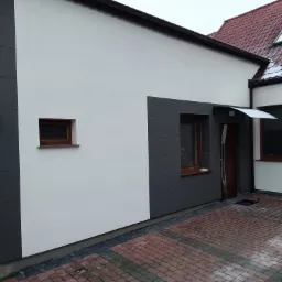 Nowoczesna elewacja domu z białym tynkiem strukturalnym i szarymi panelami, widoczne okna z drewnianymi ramami, czerwona kostka brukowa na podjeździe i drabina oparta o ścianę sąsiedniego budynku.