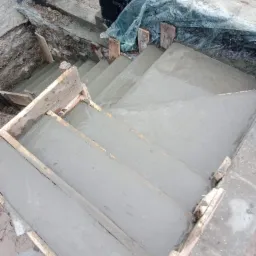 Betonowe schody w trakcie wylewania, drewniane szalunki, widoczne ślady ręcznej pracy na powierzchni betonu, surowe otoczenie budowy.