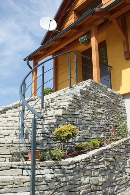 Zewnętrzne schody spiralne z szarej stali prowadzące na balkon z kamienną balustradą, żółta elewacja budynku z drewnianymi elementami, widoczna antena satelitarna i wiszące kwiaty.