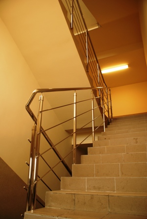 Wnętrze klatki schodowej z balustradą ze stali nierdzewnej, widok z dołu na schody wyłożone płytkami, oświetlenie jarzeniowe na suficie.