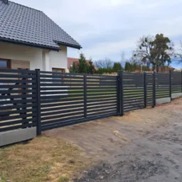 Nowoczesne, poziome ogrodzenie panelowe w kolorze antracytowym, z przesuwną bramą wjazdową. Widok na dom z szarą dachówką i podjazd.