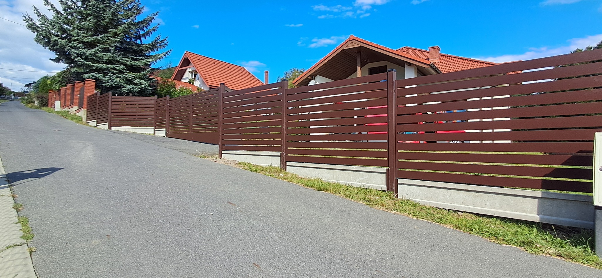 Nowoczesne, brązowe ogrodzenie panelowe z poziomych listew, zamontowane na betonowej podmurówce, wzdłuż asfaltowej drogi pod górę, w słoneczny dzień.