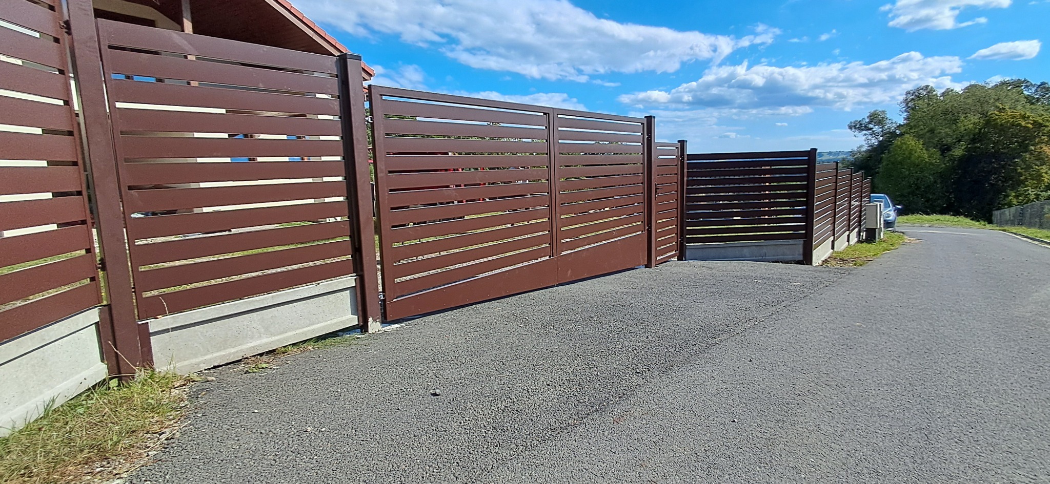 Nowoczesna, brązowa brama przesuwna z poziomymi lamelami, zamontowana na betonowej podmurówce. Widok na podjazd i fragment ogrodzenia w tle, błękitne niebo.