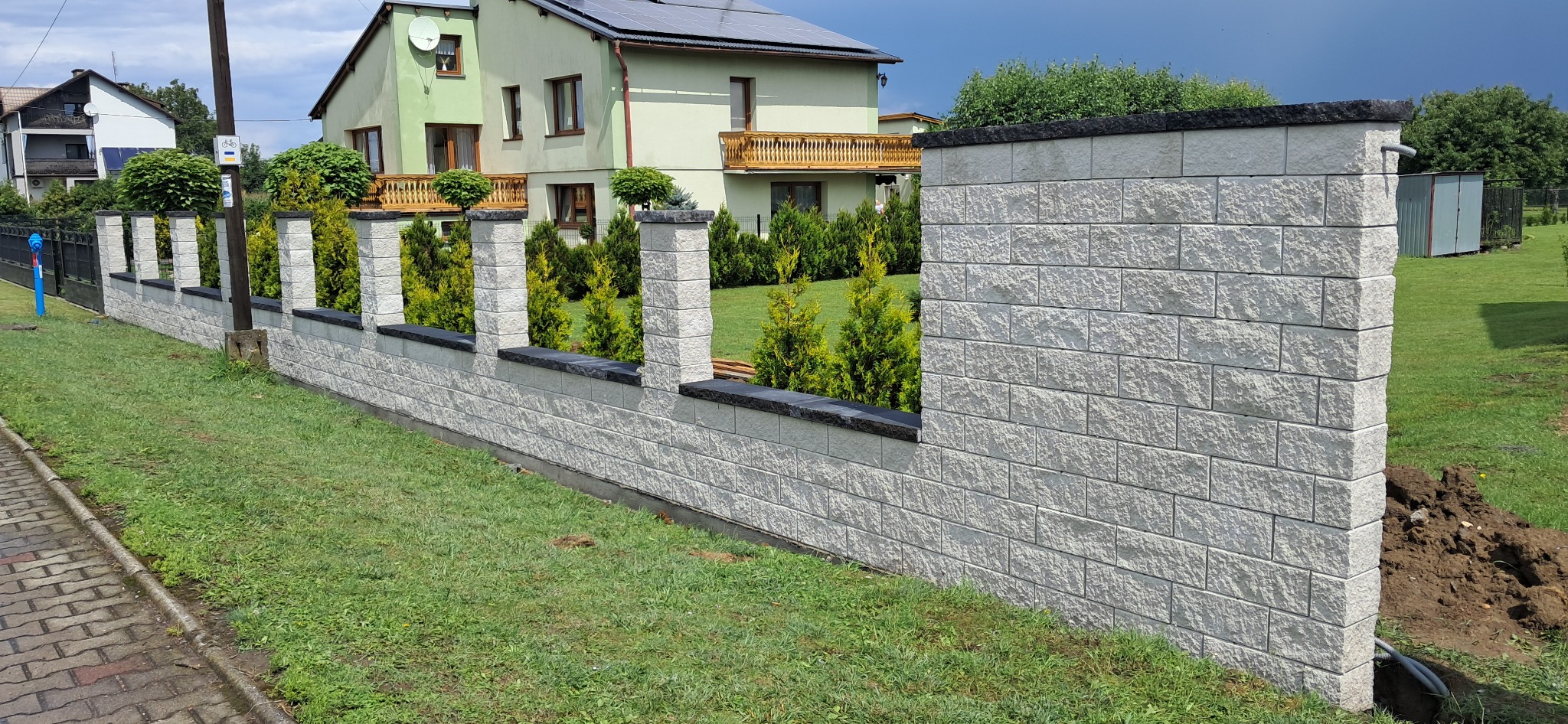 Nowoczesne ogrodzenie modułowe z jasnoszarego kamienia łupanego z czarnymi elementami wykończeniowymi, otoczone zielenią i młodymi tujami.