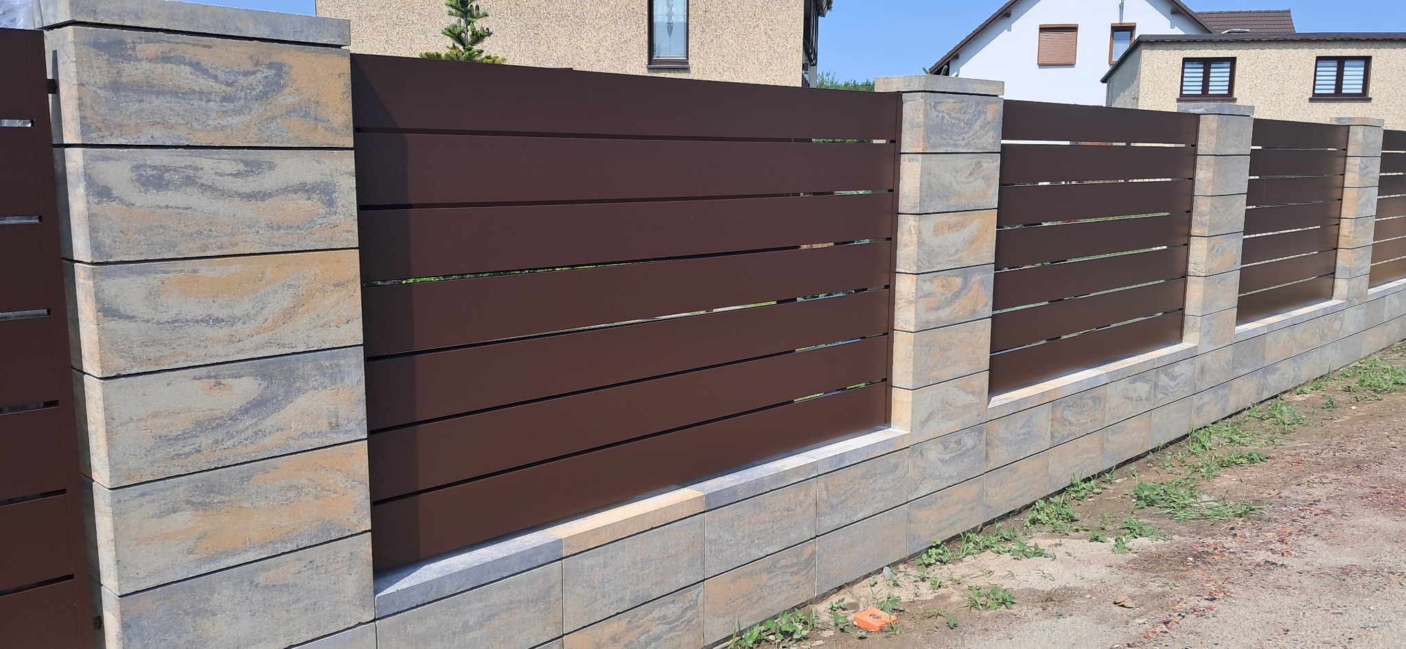 Nowoczesne ogrodzenie panelowe z brązowymi, poziomymi przęsłami i słupkami z szarej kostki o nieregularnym wzorze, na tle domów jednorodzinnych.