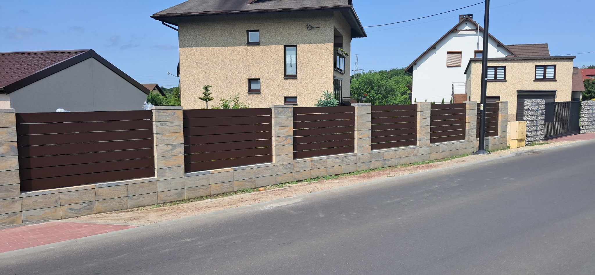 Nowoczesne ogrodzenie panelowe z brązowymi przęsłami i kamiennymi słupkami, widok z ulicy, domy w tle, słoneczny dzień.