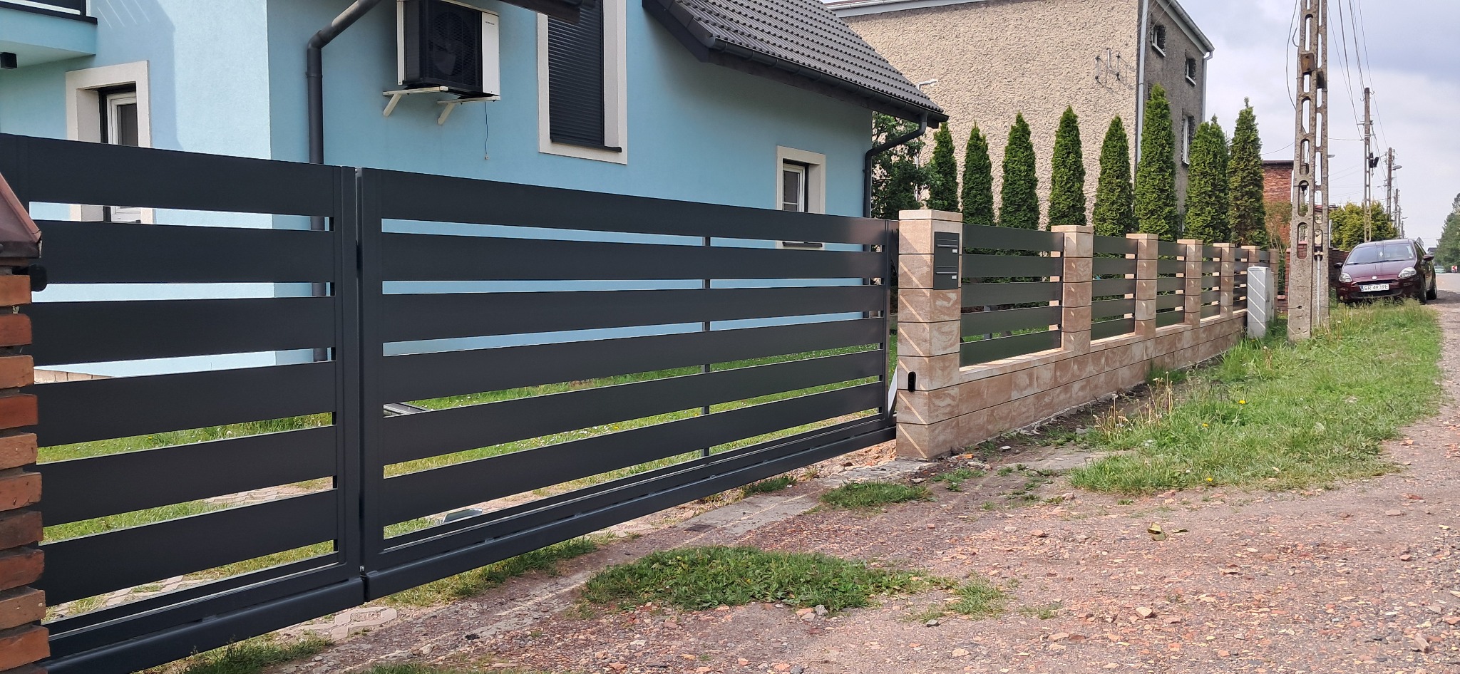 Nowoczesna, przesuwna brama w kolorze grafitowym z poziomymi panelami, zintegrowana z ogrodzeniem z ceglastymi słupkami i beżową podmurówką. Dom w tle.
