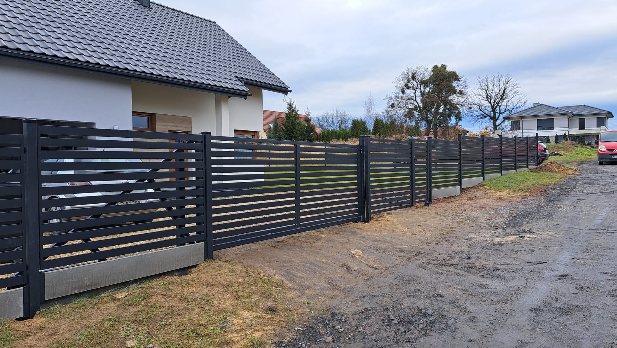 Nowoczesne, poziome ogrodzenie panelowe w kolorze antracytowym, z przesuwną bramą wjazdową. Widok na dom z szarą dachówką i podjazd.