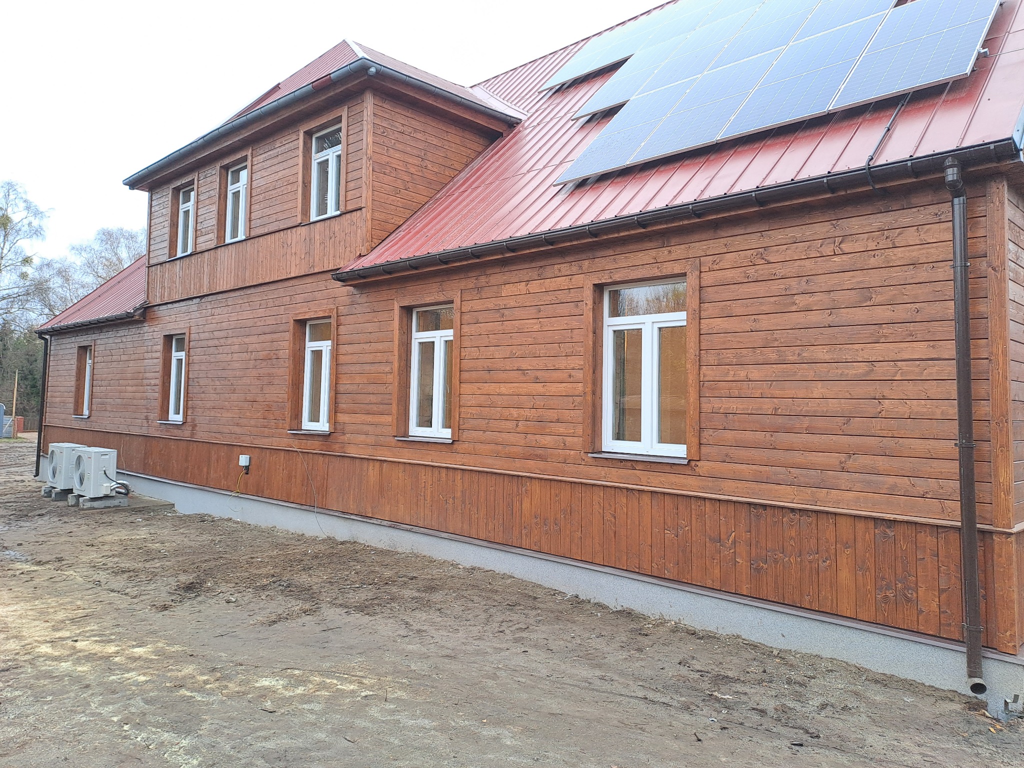 Dom z drewnianą elewacją w kolorze brązowym, czerwony dach z panelami słonecznymi, widoczne dwie jednostki klimatyzacyjne na zewnątrz budynku.