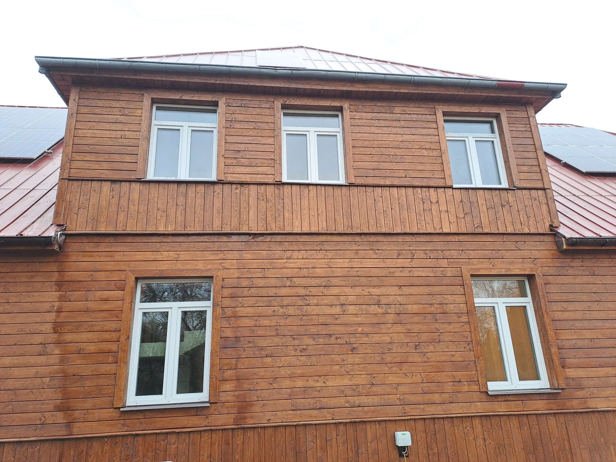 Drewniana elewacja domu z białymi oknami i czerwonym dachem, na którym zamontowane są panele słoneczne.