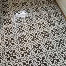 Wzór podłogi z płytek ceramicznych w stylu retro, czarno-biały motyw kwiatowy z elementami geometrycznymi, widoczna fuga i fragment białej listwy przypodłogowej.