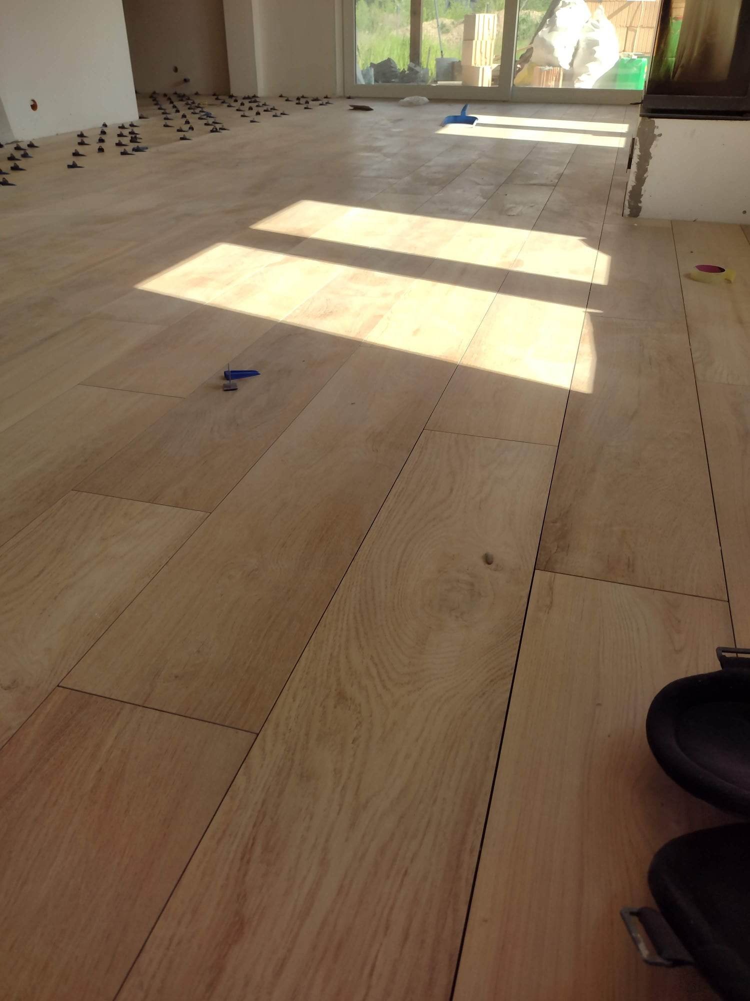 Układanie podłogi z płytek imitujących drewno, z użyciem niebieskich klinów poziomujących, w jasnym pomieszczeniu z dużym oknem, w tle widoczne zapasy płytek.