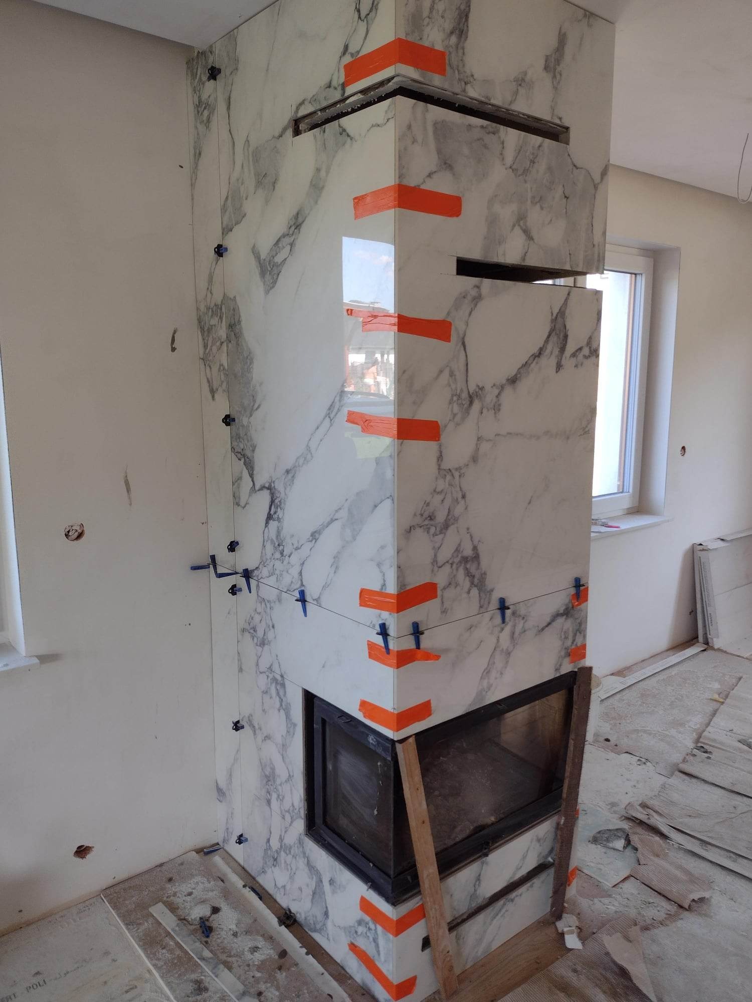 Wykończenie kominka płytkami imitującymi marmur, widoczne niebieskie krzyżyki dystansowe i pomarańczowa taśma zabezpieczająca narożniki, surowy stan wnętrza.