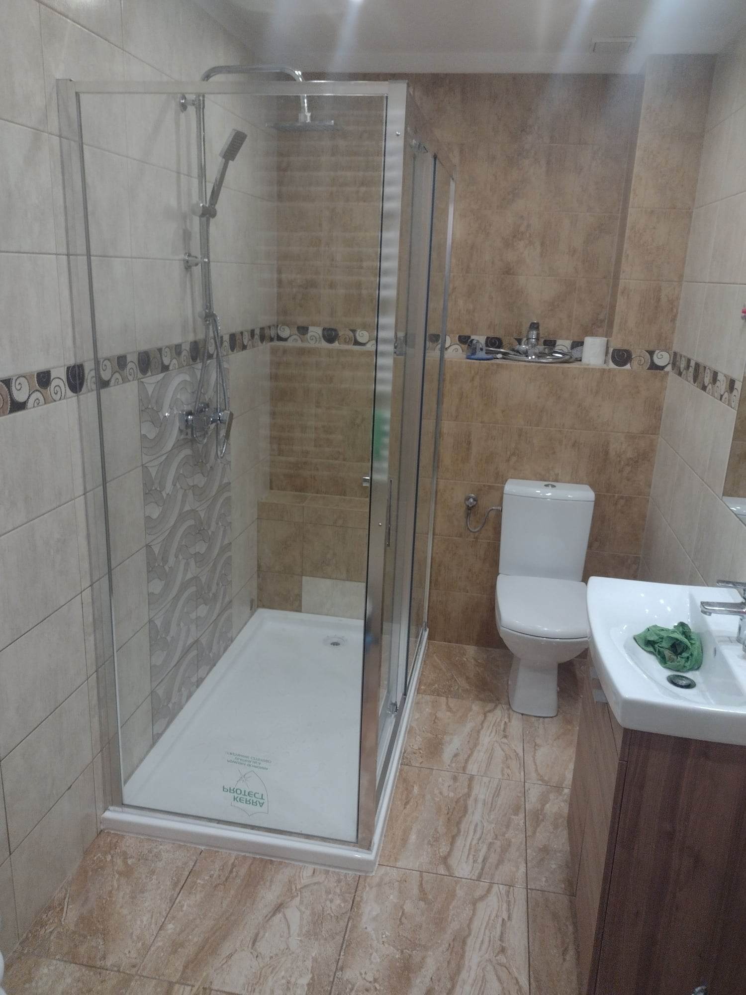 Wykończona łazienka z kabiną prysznicową z przezroczystego szkła, jasnymi płytkami z dekoracyjnym pasem i beżową mozaiką, białą toaletą i umywalką z brązową szafką.
