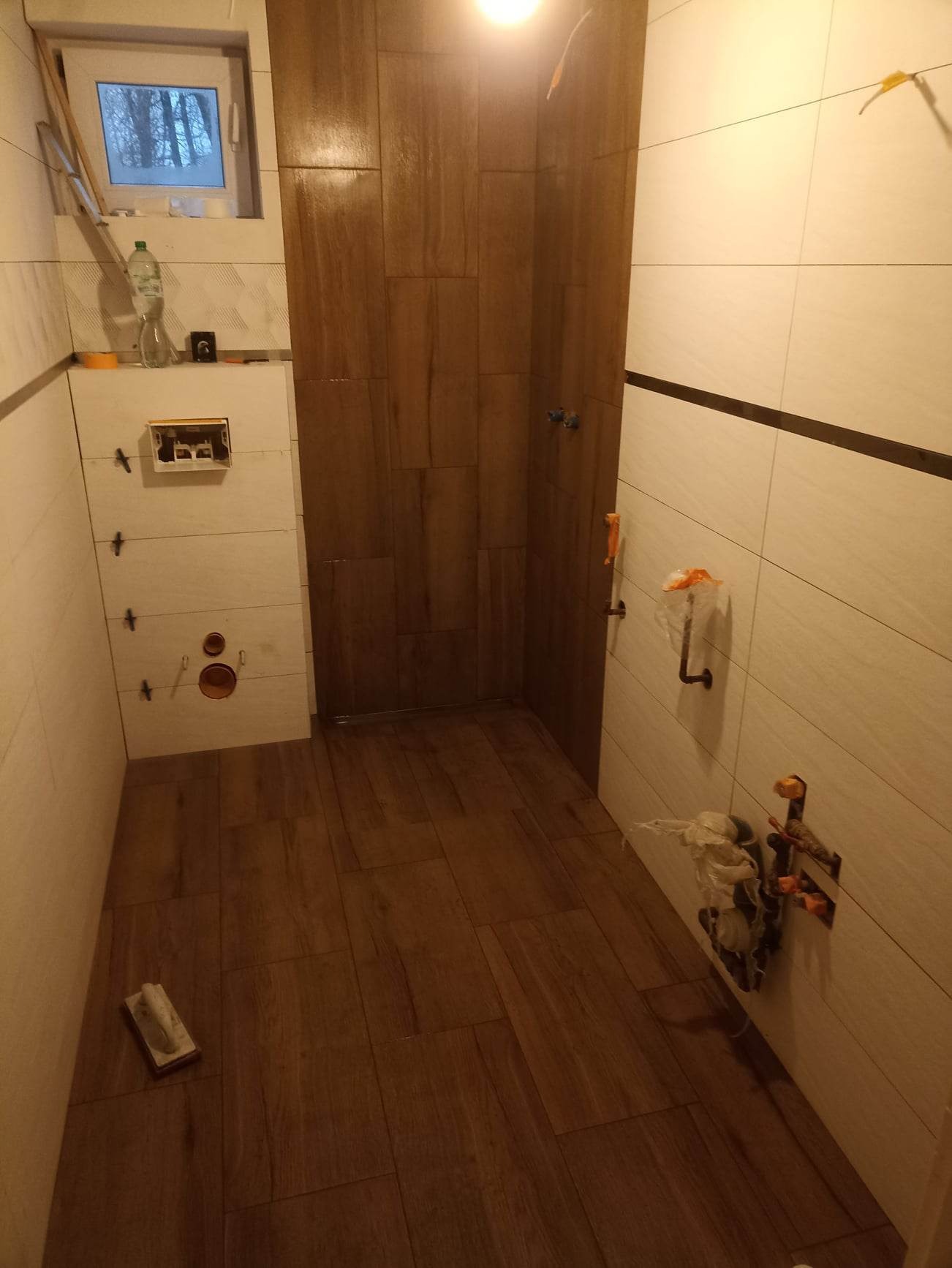 Wnętrze łazienki w trakcie remontu, widoczne płytki imitujące drewno na podłodze i ścianie prysznica, białe płytki na pozostałych ścianach z czarnym pasem dekoracyjnym, wystające rury i elementy...