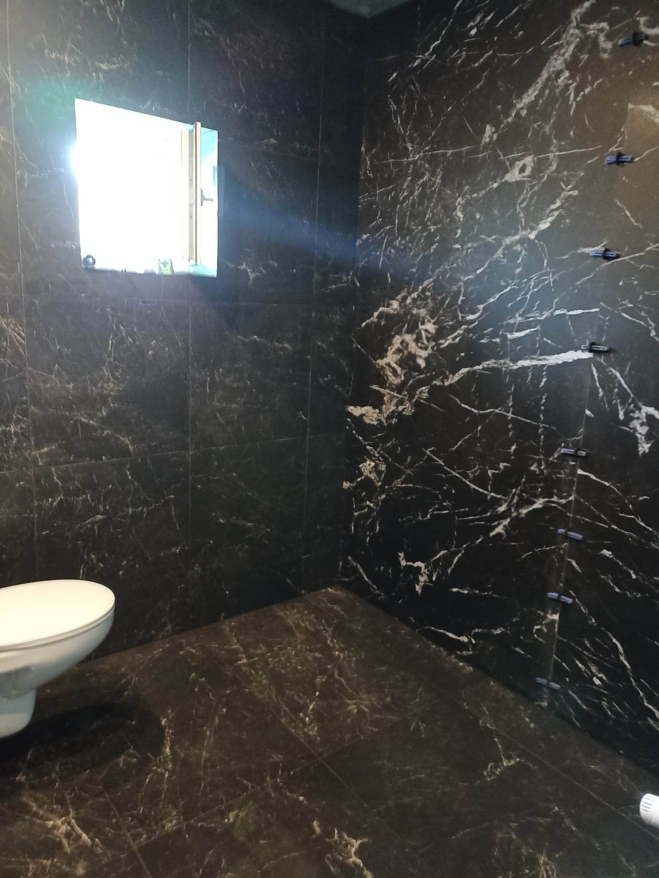 Ciemna łazienka z marmurowymi płytkami, widoczne krzyżyki dystansowe na ścianie i podwieszana toaleta.