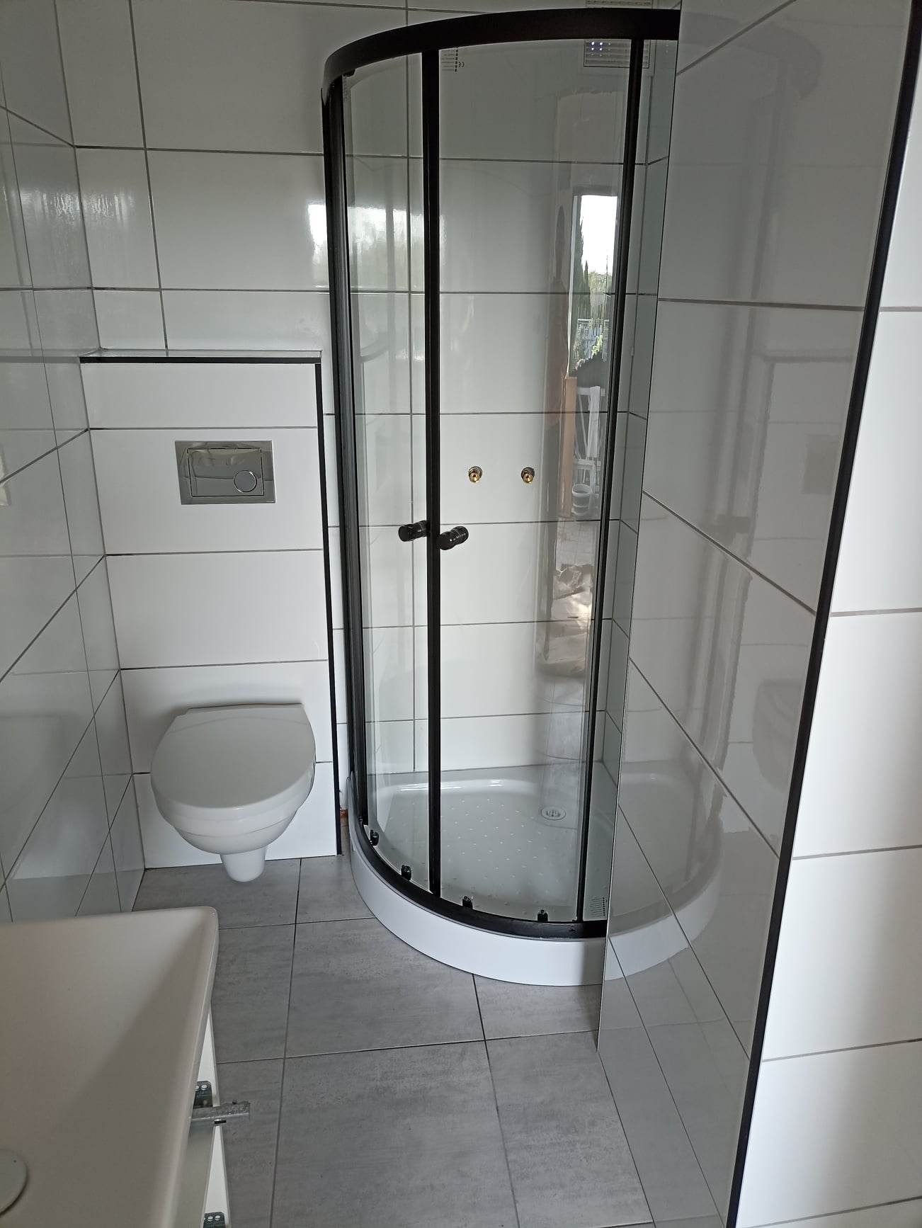 Wykończona łazienka z kabiną prysznicową z czarnymi ramami, wiszącą toaletą z chromowanym przyciskiem spłukiwania i szarymi płytkami podłogowymi.