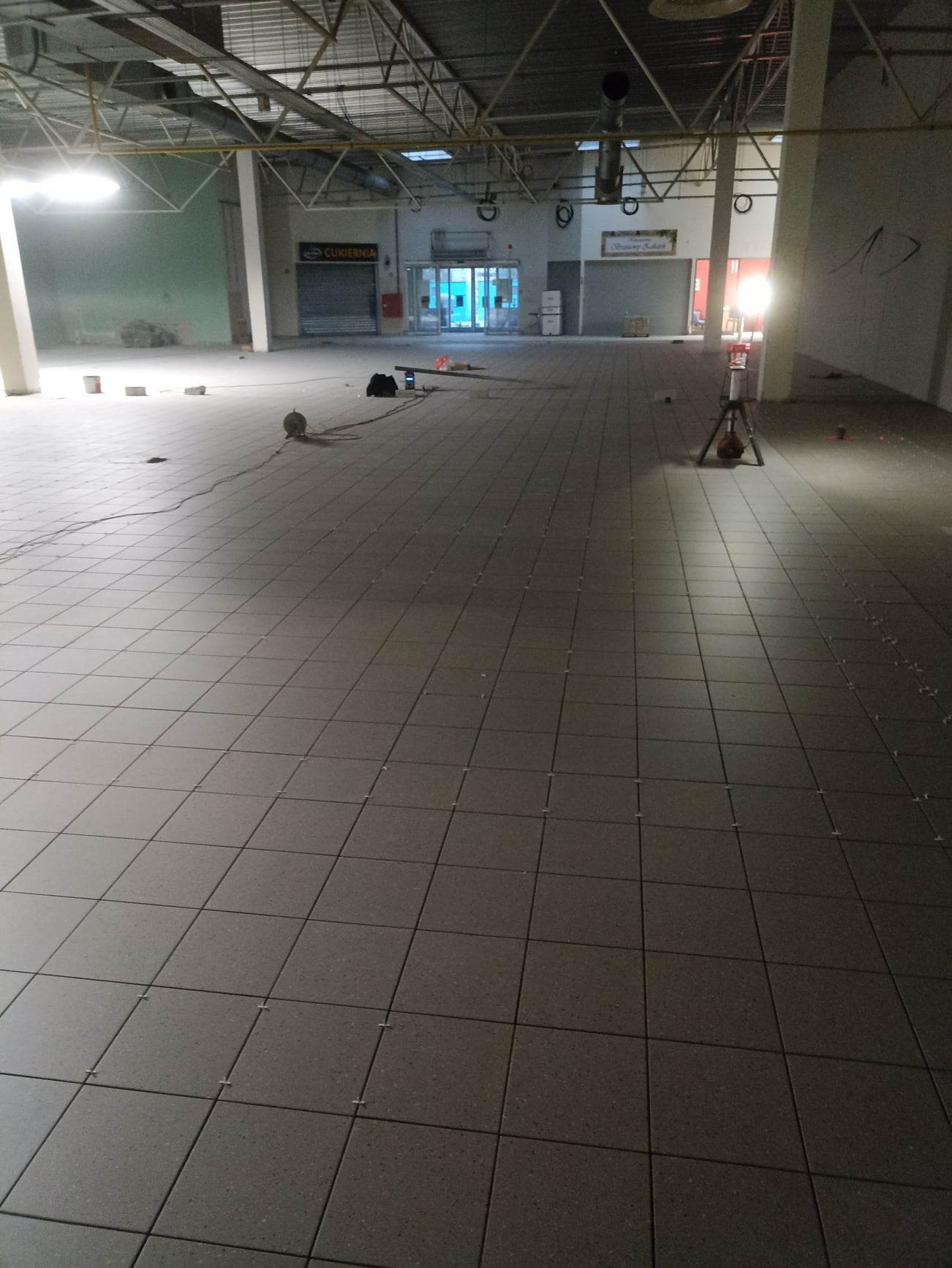 Nowo ułożona, szara, kwadratowa podłoga z płytek w przestronnym, pustym lokalu handlowym. Widoczne krzyżyki dystansowe między płytkami, lampa budowlana na statywie, narzędzia i przewody w tle.