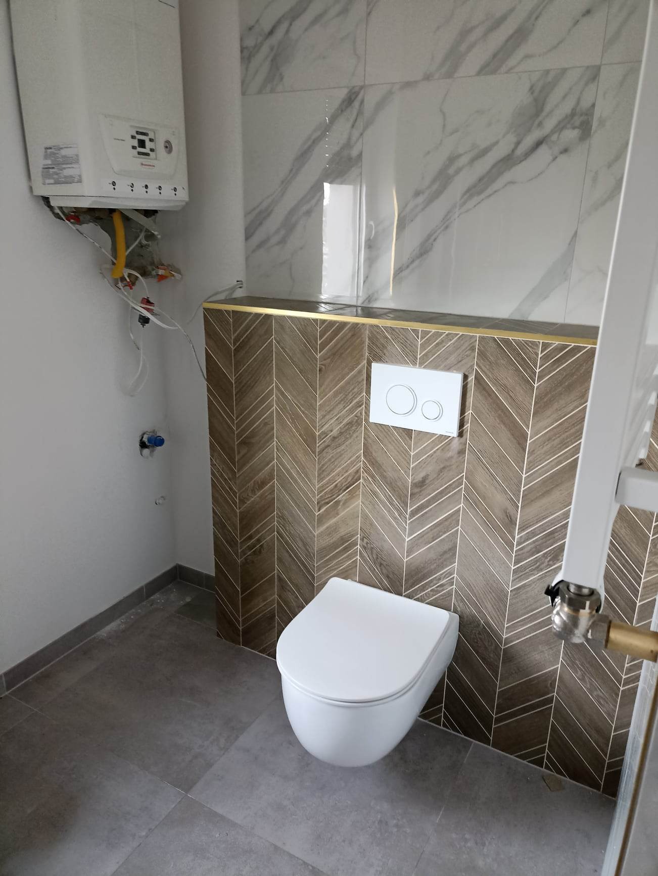 Nowoczesna toaleta podwieszana z brązową, wzorzystą okładziną ścienną, marmurowe płytki na górze, wiszący bojler z widocznymi instalacjami, szare płytki podłogowe.