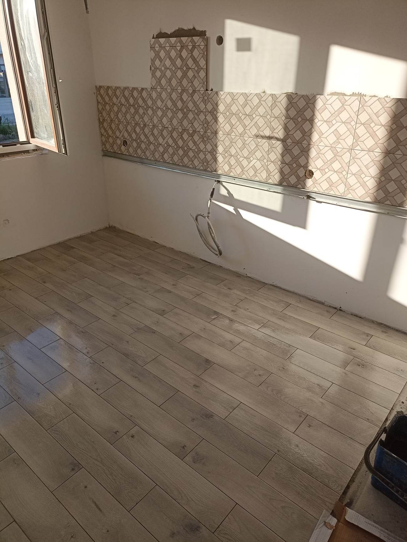Wnętrze w trakcie remontu: ułożona podłoga z paneli imitujących drewno, fragment ściany z częściowo położonymi płytkami w geometryczny wzór, widoczna metalowa listwa montażowa i wystające kable.
