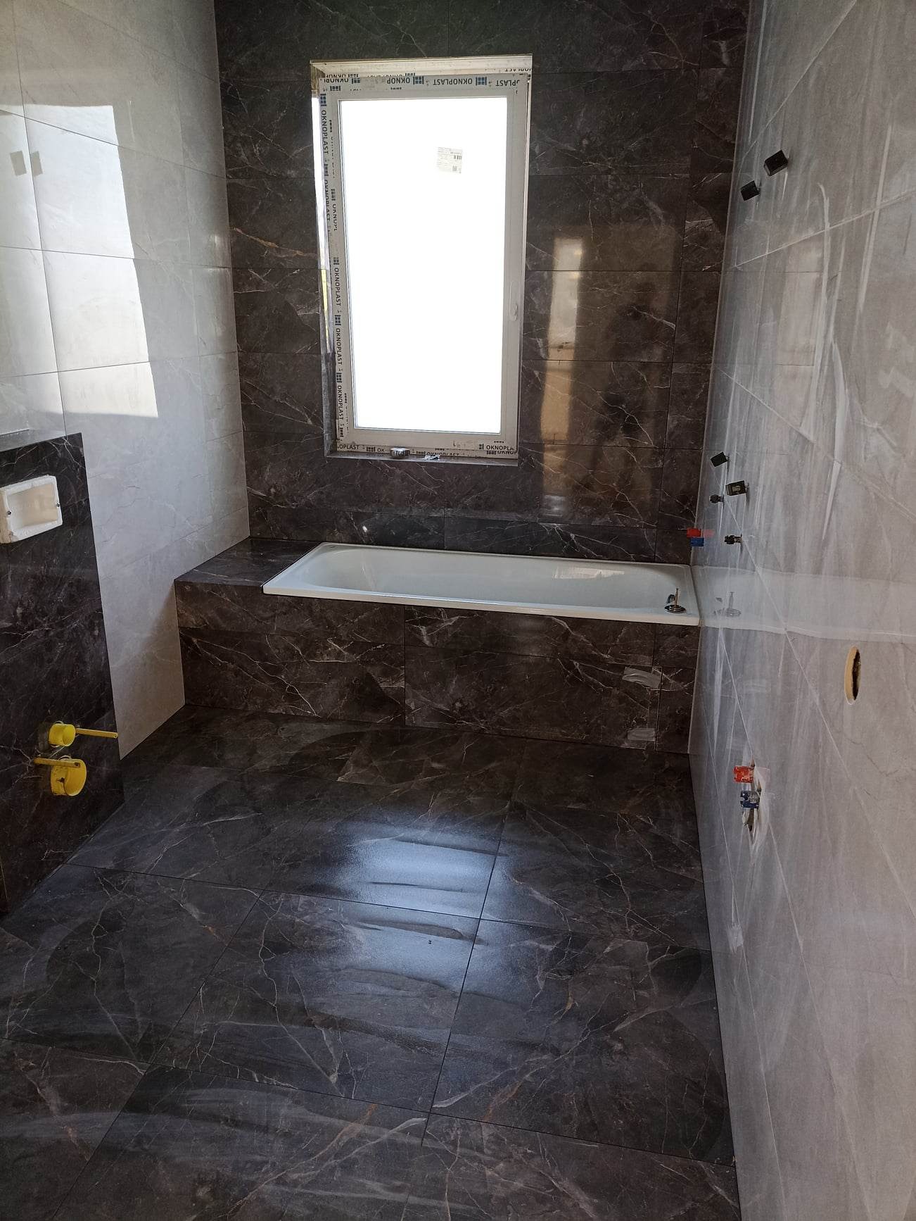 Nowoczesna łazienka z ciemnymi, marmurkowymi płytkami na podłodze i ścianach, biała wanna pod oknem, widoczne instalacje wodne w ścianie, surowe wykończenie.