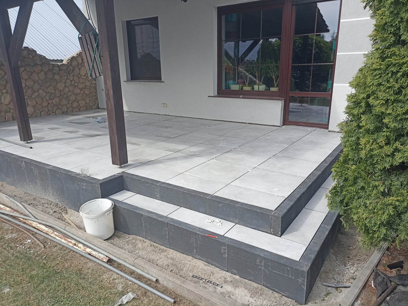 Jasnoszare płytki gresowe ułożone na tarasie z dwoma stopniami, z widocznymi krzyżykami dystansowymi, białe ściany budynku z brązowymi oknami, z boku iglak, wiadro i rury na ziemi.