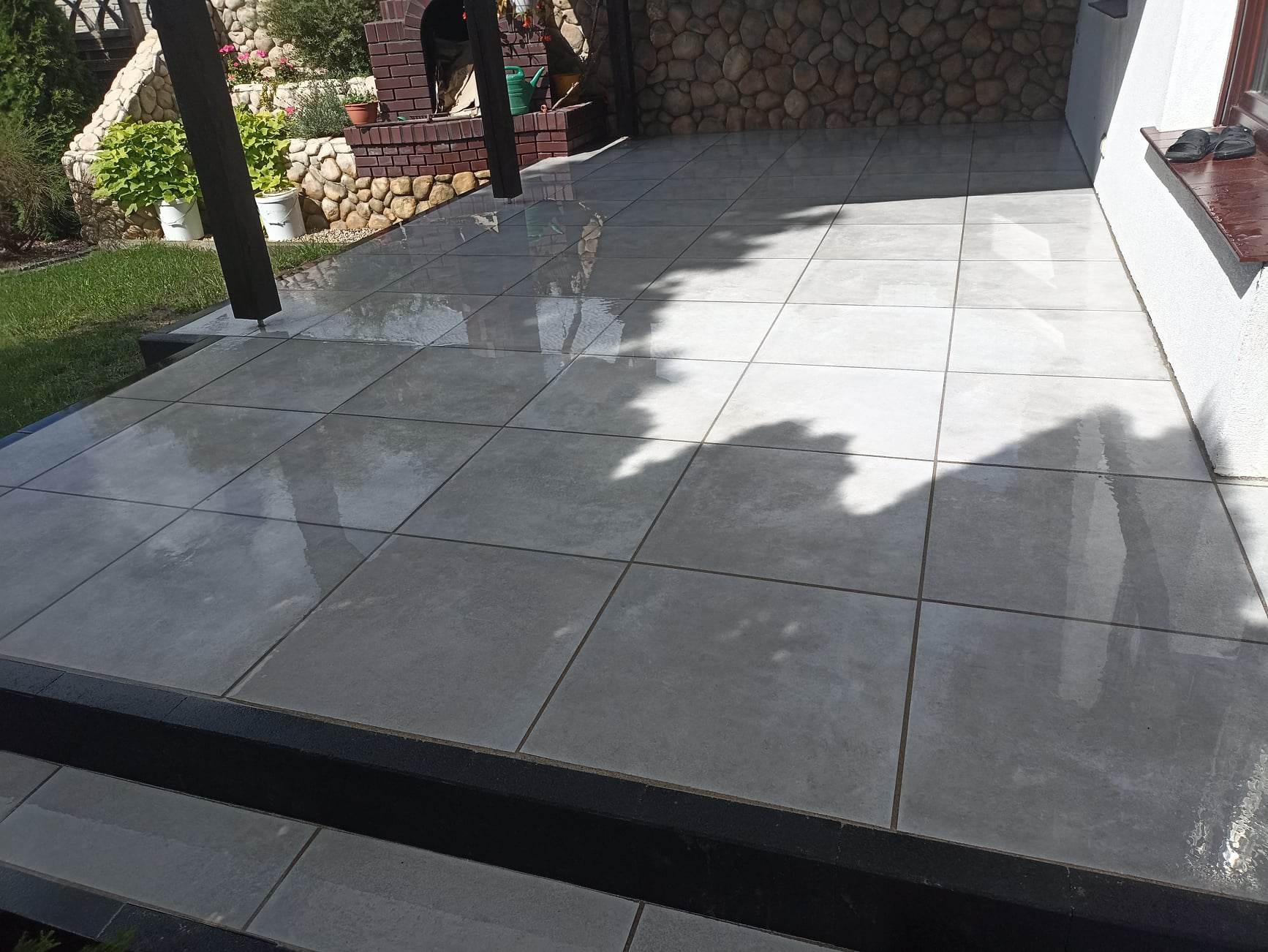 Świeżo ułożona, jasnoszara, kwadratowa terakota na tarasie, częściowo mokra od niedawnego mycia, z widocznym fragmentem schodka z czarnego kamienia i elementami ogrodu w tle.