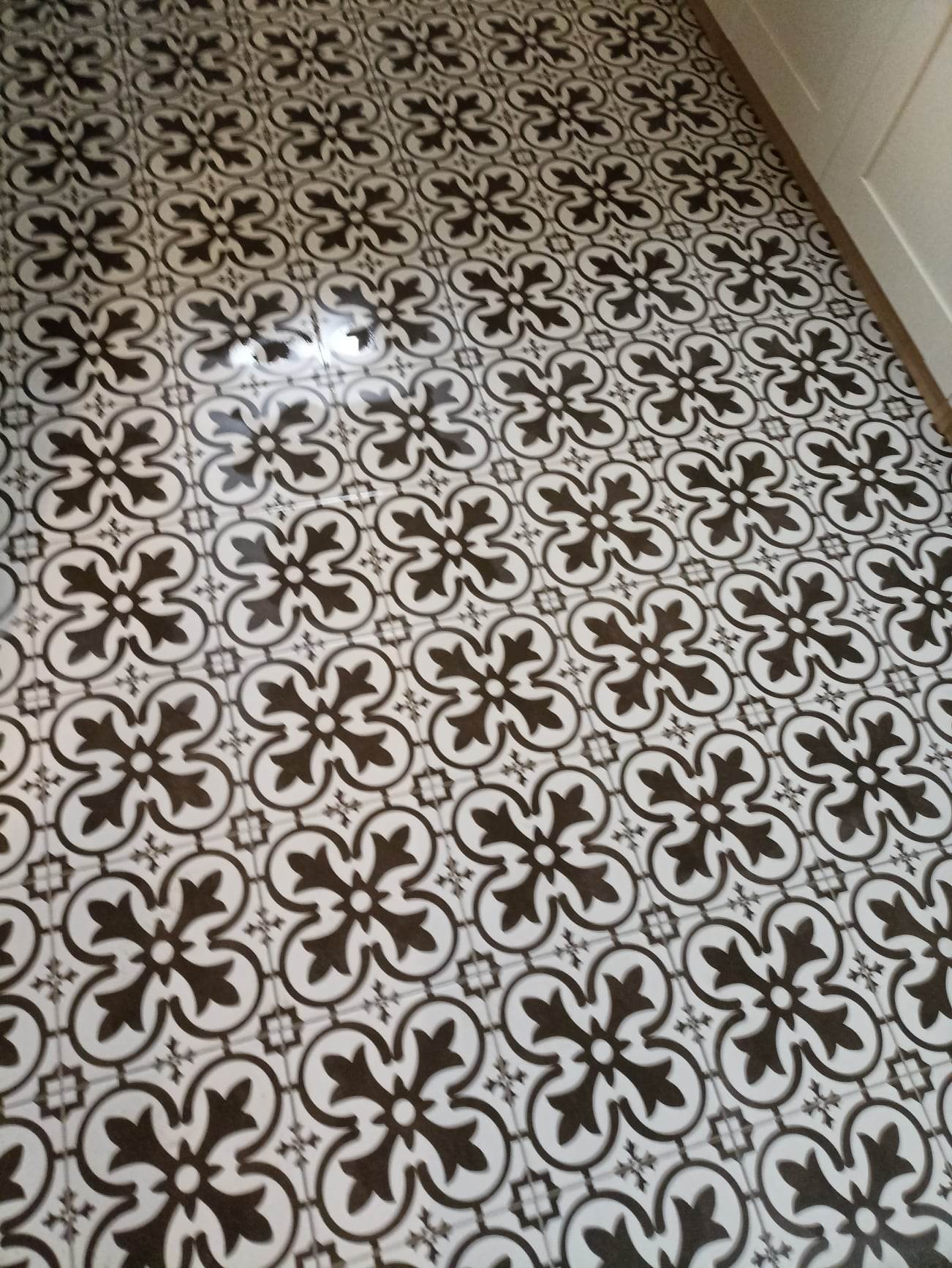 Wzór podłogi z płytek ceramicznych w stylu retro, czarno-biały motyw kwiatowy z elementami geometrycznymi, widoczna fuga i fragment białej listwy przypodłogowej.