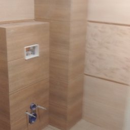 Styl-Kam - Obudowa stelaża WC z płytek imitujących drewno, widoczne przyłącza wodne i otwór rewizyjny, jasne płytki podłogowe i ścienne, fragment wykończonej łazienki.