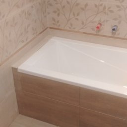 Styl-Kam - Wanna obudowana brązowymi płytkami, ściany wyłożone beżowymi płytkami z motywem liści, widoczne przyłącza wodne.