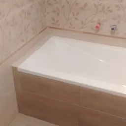 Wanna obudowana brązowymi płytkami, ściany wyłożone beżowymi płytkami z motywem liści, widoczne przyłącza wodne.