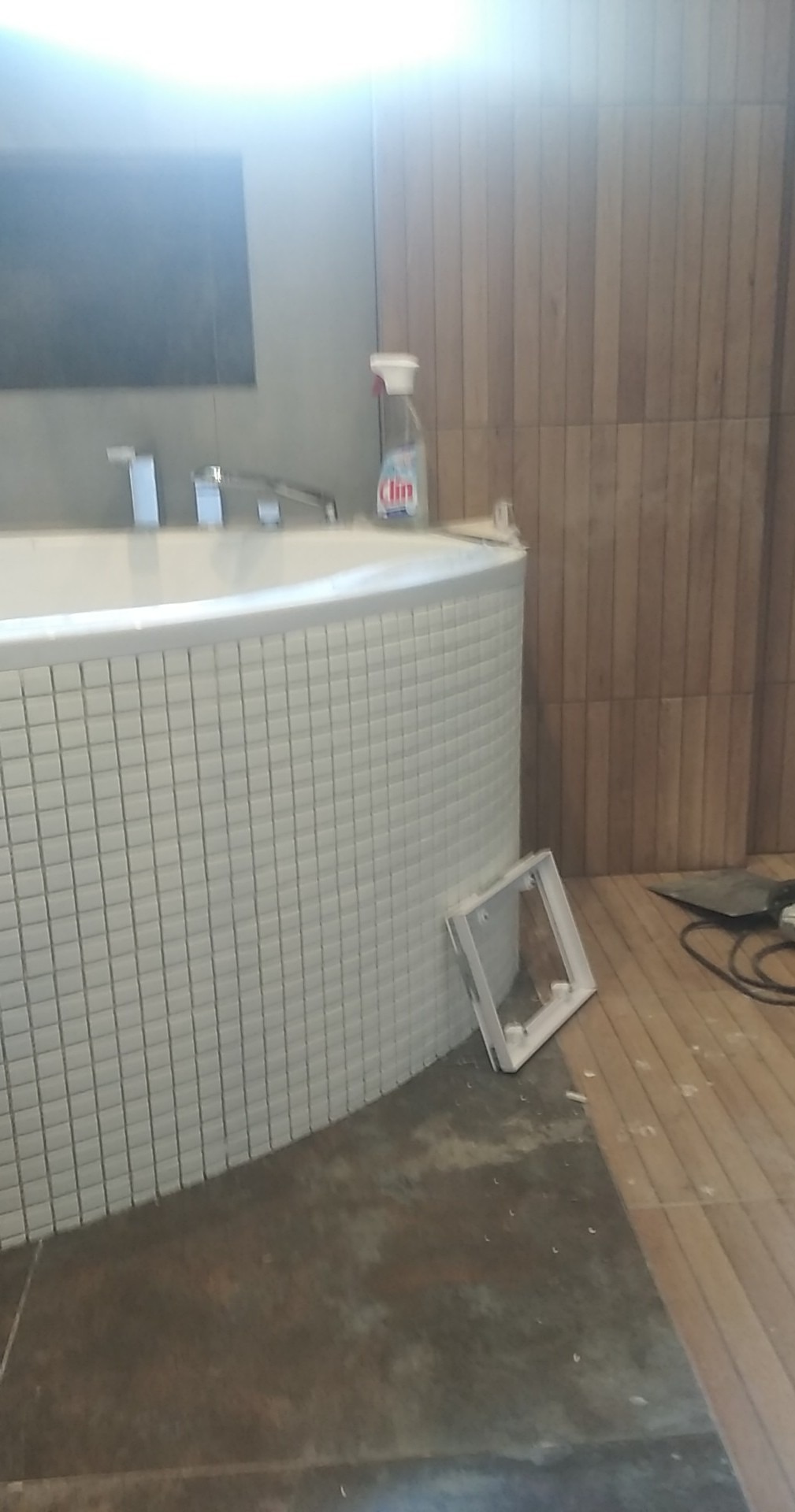 Okrągła wanna obłożona białą mozaiką w trakcie prac wykończeniowych, widoczne niedociągnięcia na podłodze i ścianie, oraz narzędzia i akcesoria do sprzątania.