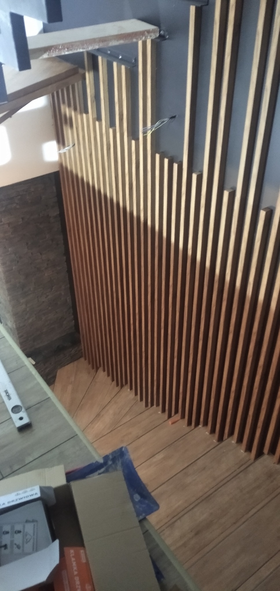 Drewniane lamele na ścianie klatki schodowej, widoczne przewody elektryczne, schody z drewnopodobnych płytek, poziomica i kartonowe opakowania w dolnej części kadru.