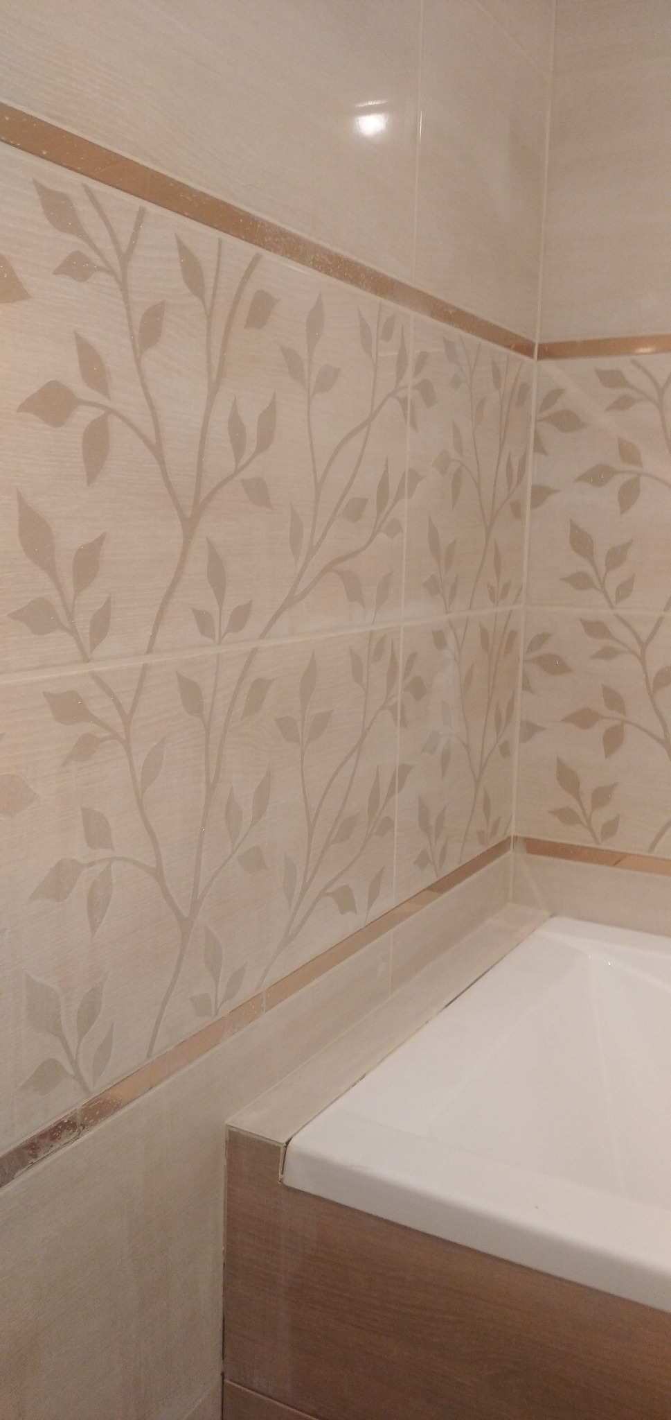 Beżowe płytki ceramiczne z delikatnym wzorem liści na ścianie nad wanną, wykończone brązowymi listwami.