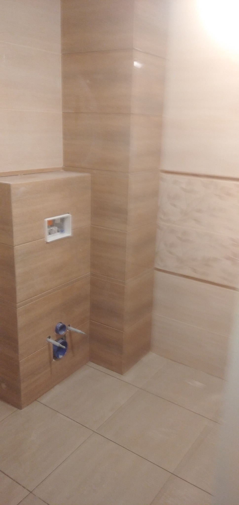 Obudowa stelaża WC z płytek imitujących drewno, widoczne przyłącza wodne i otwór rewizyjny, jasne płytki podłogowe i ścienne, fragment wykończonej łazienki.