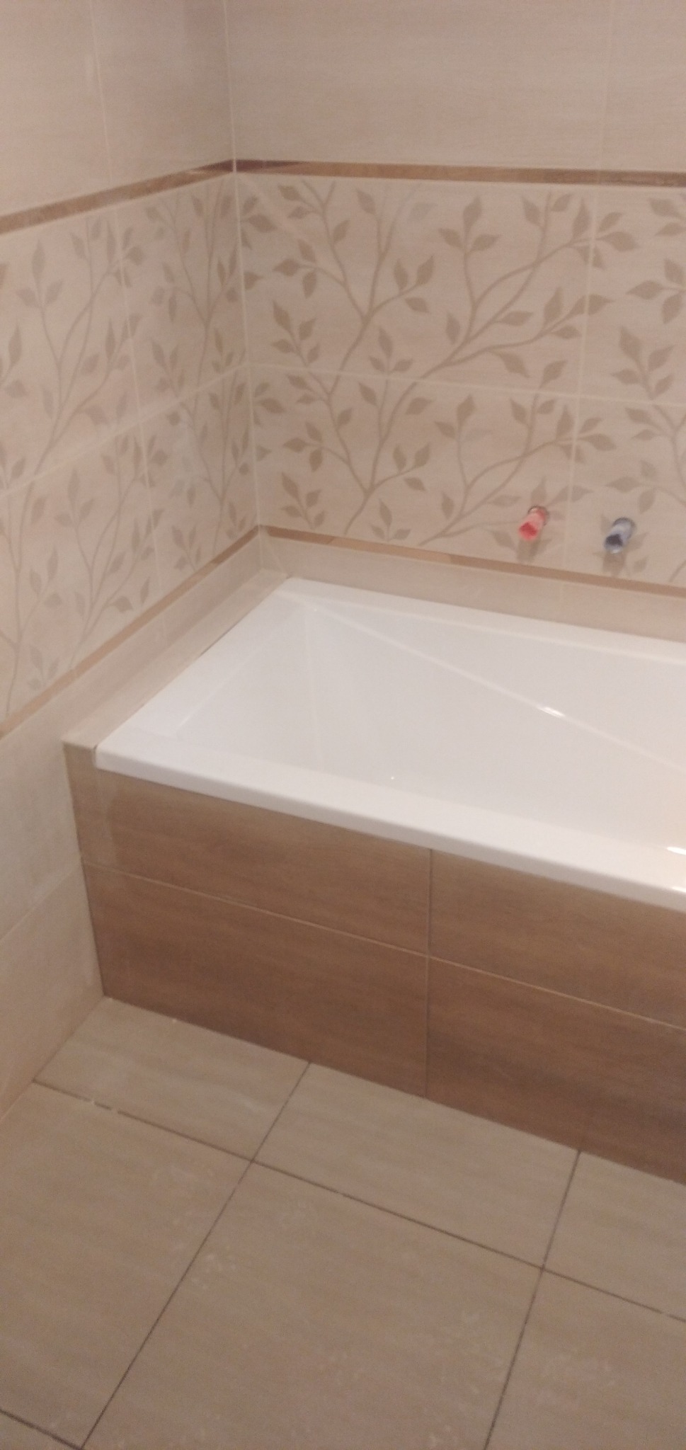 Wanna obudowana brązowymi płytkami, ściany wyłożone beżowymi płytkami z motywem liści, widoczne przyłącza wodne.