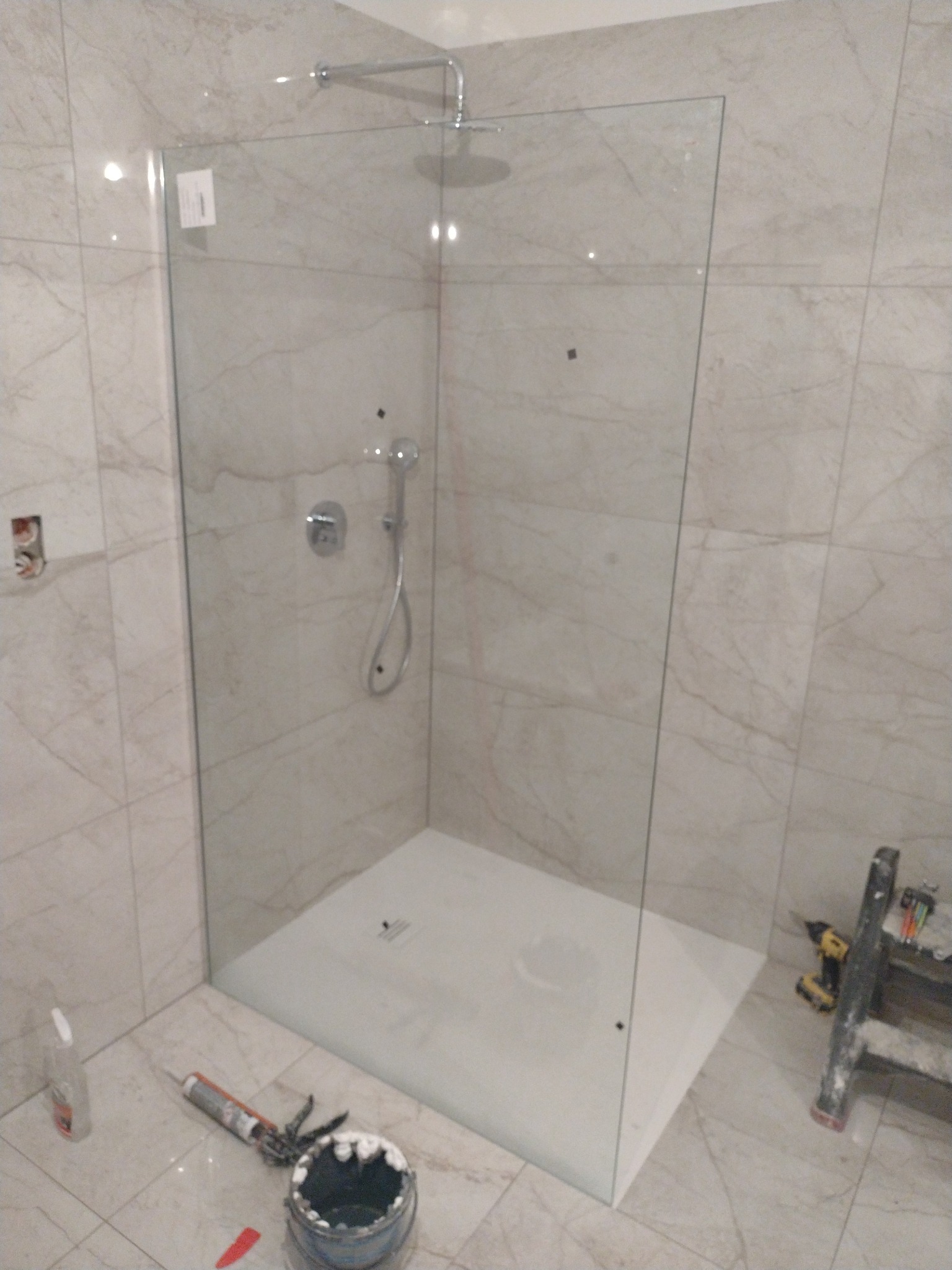 Nowoczesna kabina prysznicowa typu walk-in z chromowaną armaturą i deszczownicą, zamontowana w łazience wyłożonej płytkami imitującymi marmur, widoczne akcesoria montażowe i drabina.