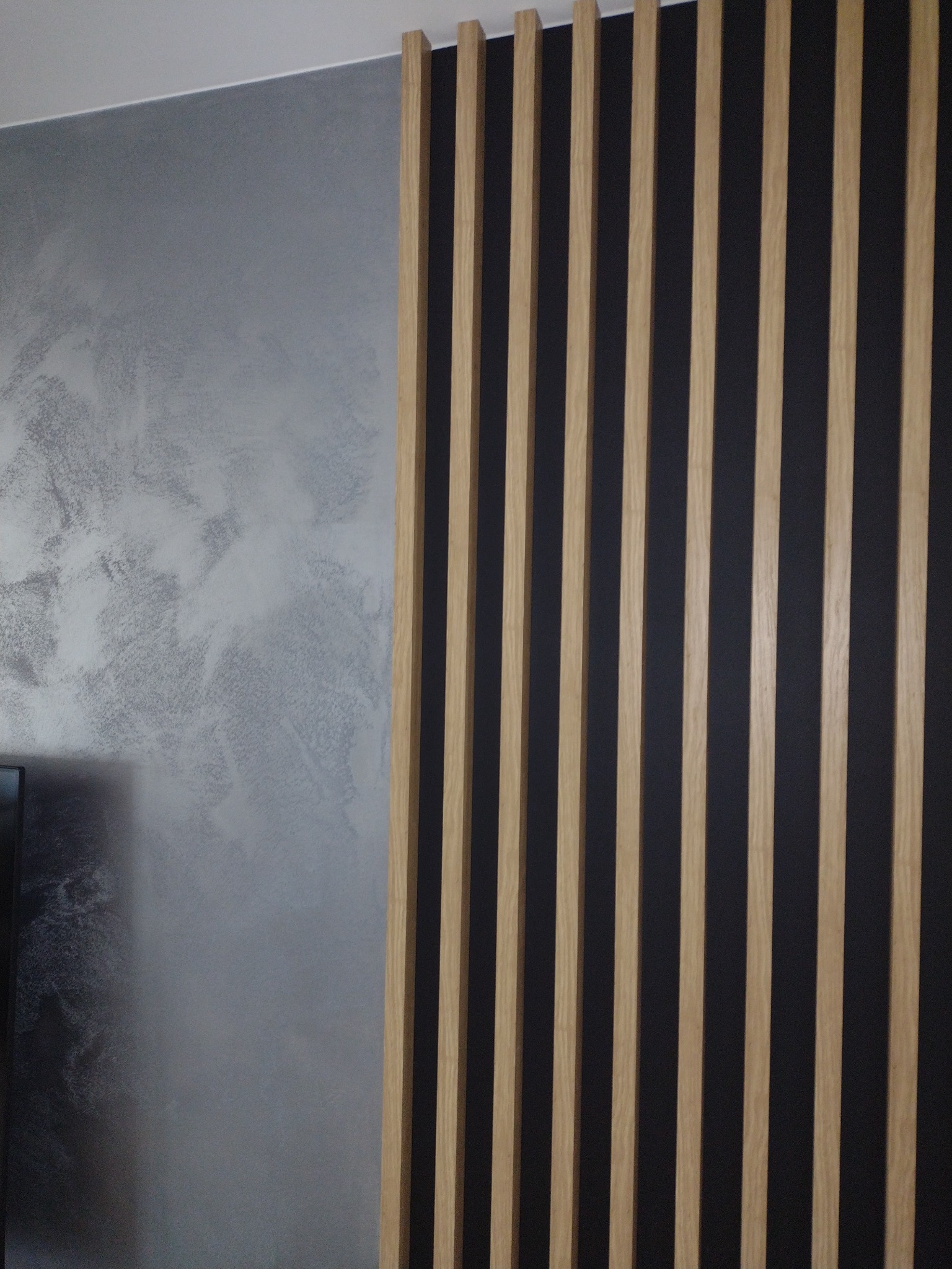 Dekoracyjna ściana z pionowych, drewnianych listew na tle ciemnej ściany oraz ściany z efektem dekoracyjnym imitującym chmurę.