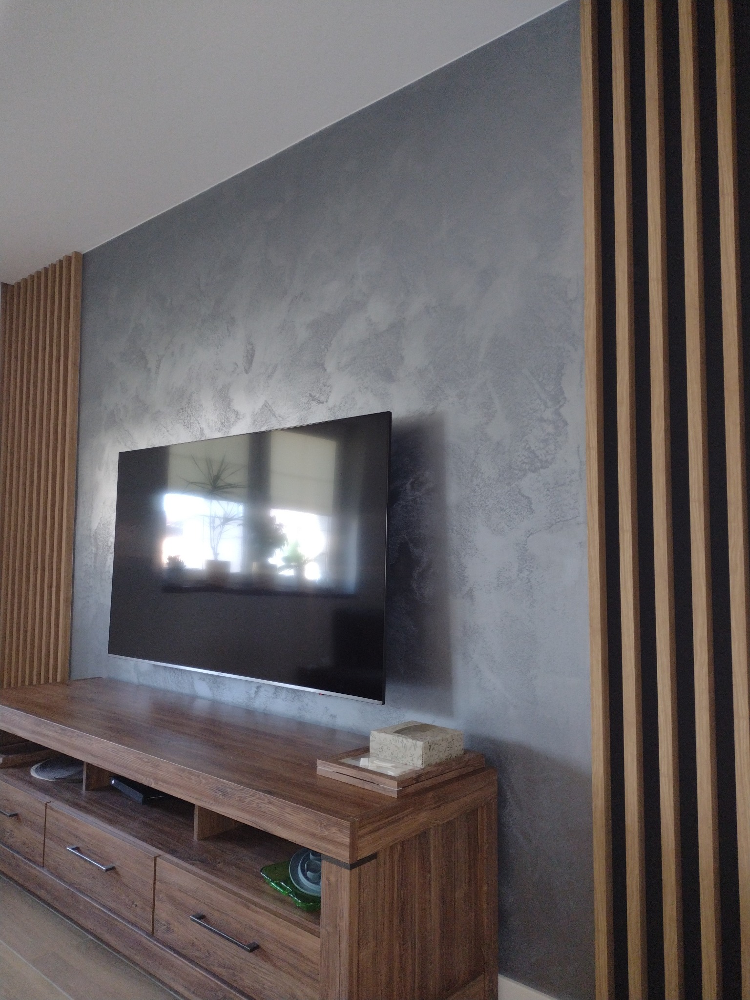 Ściana w salonie wykończona dekoracyjnym tynkiem w odcieniach szarości, z drewnianymi lamelami po bokach, pod telewizorem szafka RTV z drewna w ciepłym odcieniu.