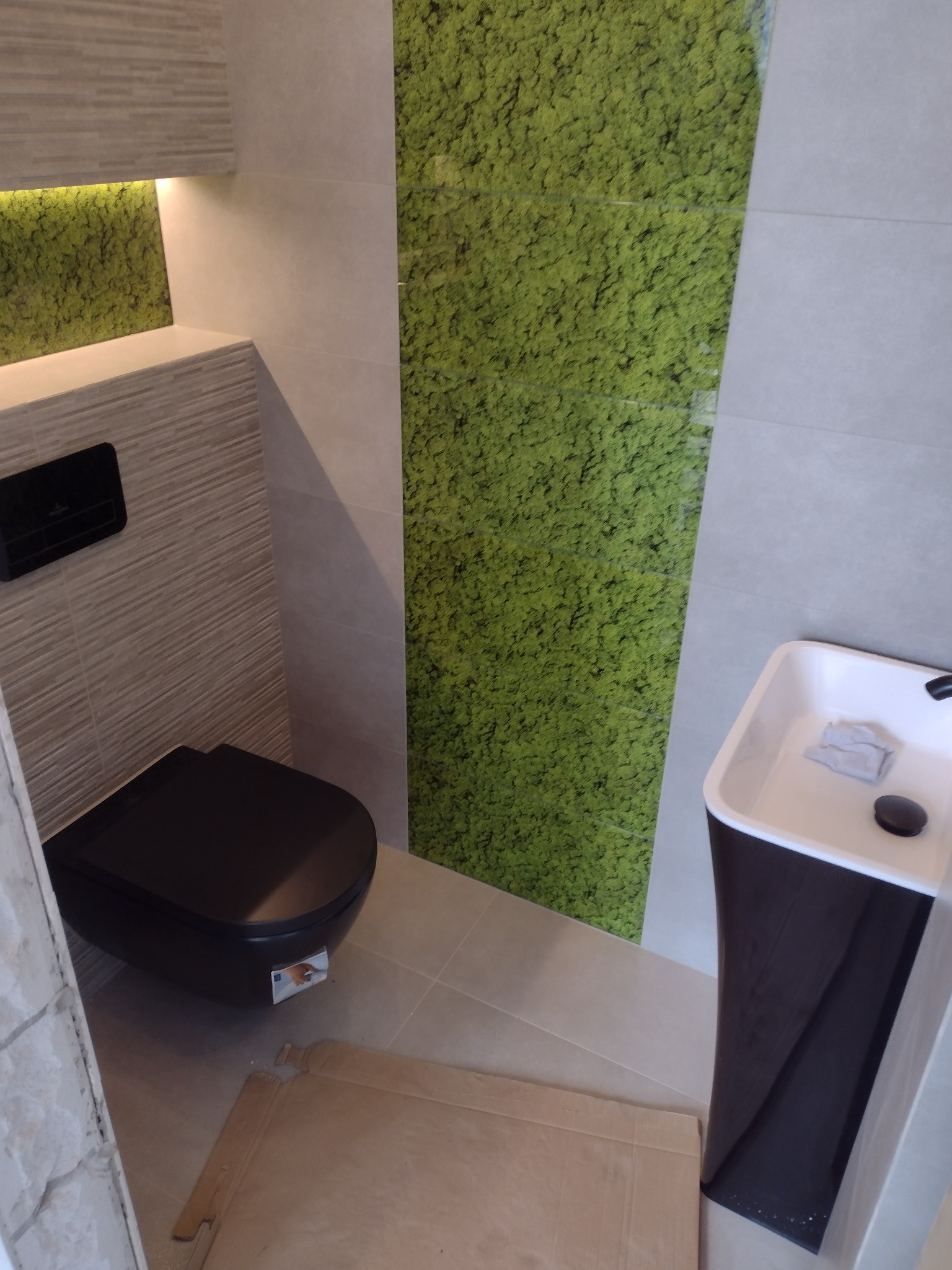 Nowoczesna łazienka z czarną toaletą podwieszaną, umywalką na postumencie i ścianą pokrytą panelami imitującymi mech.
